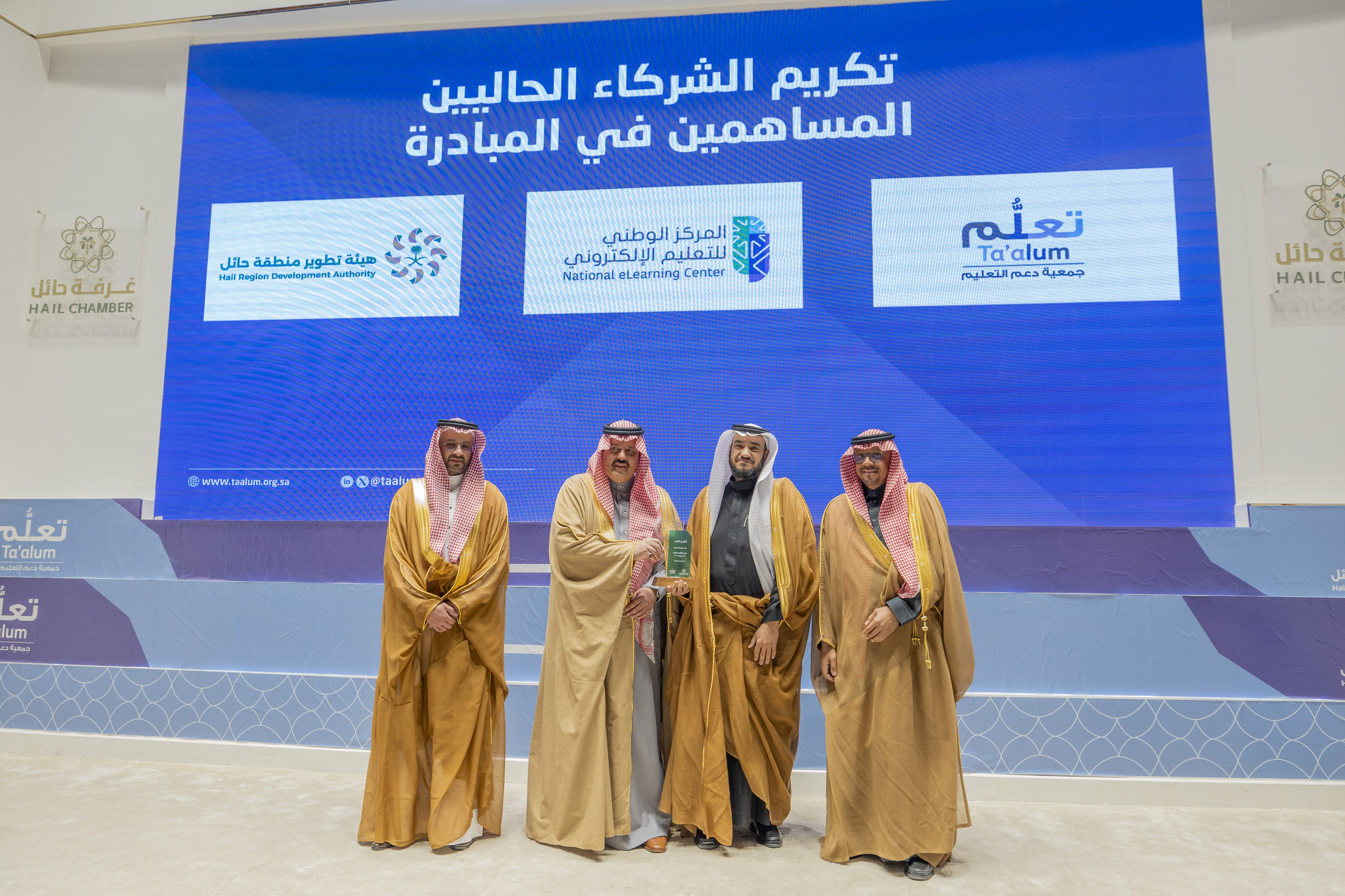 صورة لتكريم المركز الوطني للتعليم الإلكتروني كشريك في مبادرة "تعلم" برعاية صاحب السمو الملكي أمير منطقة حائل، حيث تعكس الصورة صاحب السمو الملكي أمير منطقة حائل إلى جانب 3 رجال، بالإضافة إلى شاشة بالخلف تعكس شعار جمعية دعم التعليم، المركز الوطني للتعليم الإلكتروني، وشعار هيئة تطوير منطقة حائل.