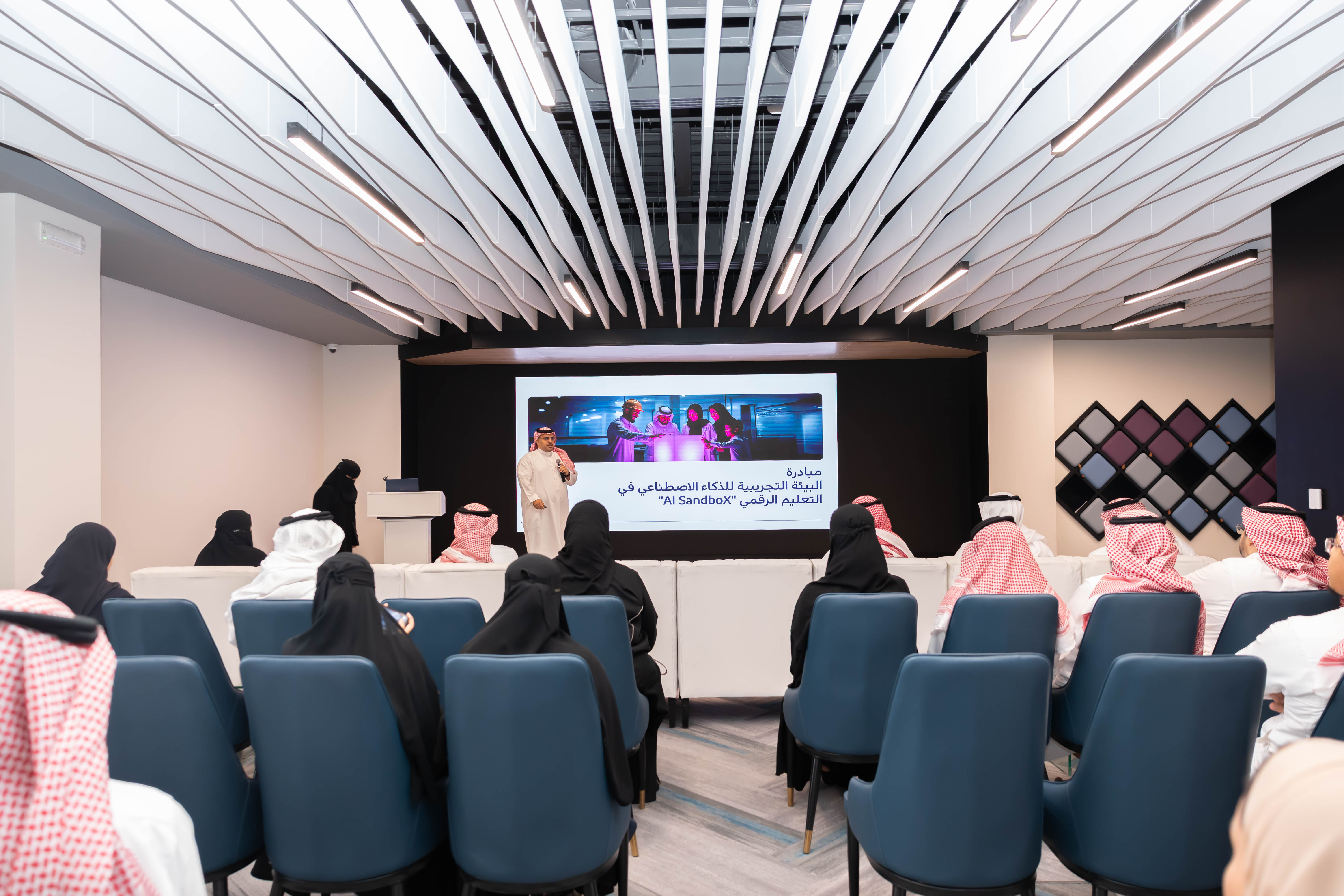 أحد منسوبي المركز، يقف أمام شاشة عرض، يستعرض عرض مرئي عن مبادرة البيئة التجريبية للذكاء الاصطناعي في التعليم الرقمي "AI SandboX" ويتضح عدد من الحضور من الرجال والنساء.